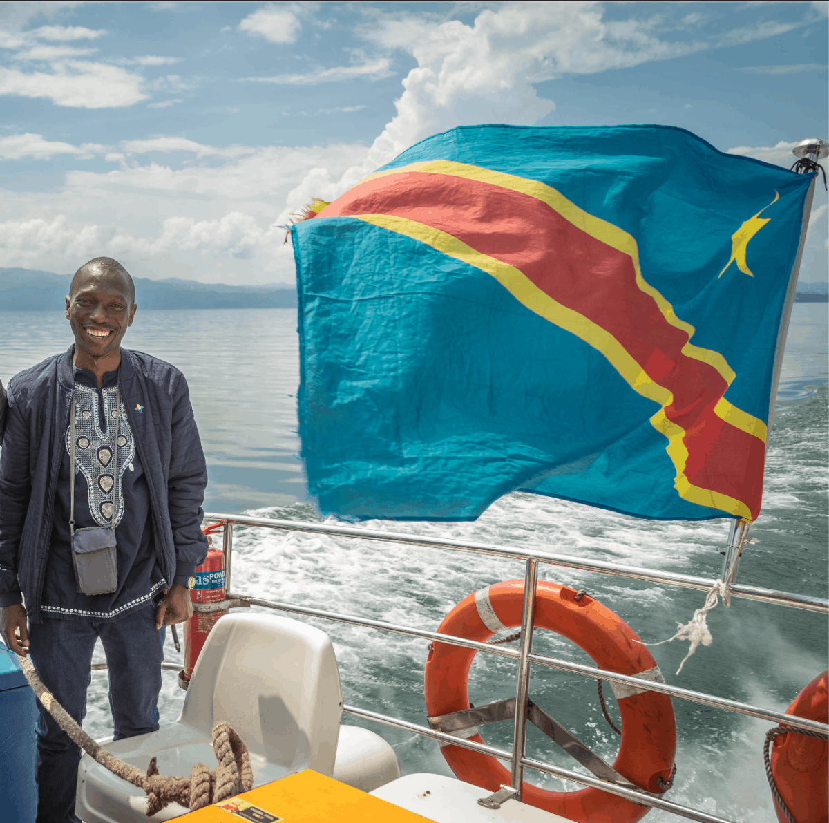 Hermann Aho, in a jacket and embroidered black shirt, smiles for the camera on a sunny day on the Lake Kivu ferry, with the DRC flag. Photo: Jon Atwell