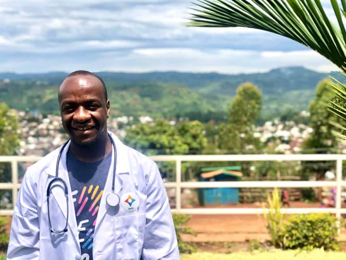 Dr. Johny Muhindo, in a white lab coat with a stethoscope, is photographed outdoors in Bukavu, DRC.
