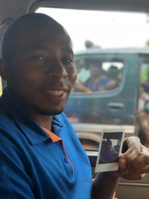 Dario Tabaro Mutarushwa, a driver, shows a polaroid photograph of himself in the driver's seat of a vehicle.