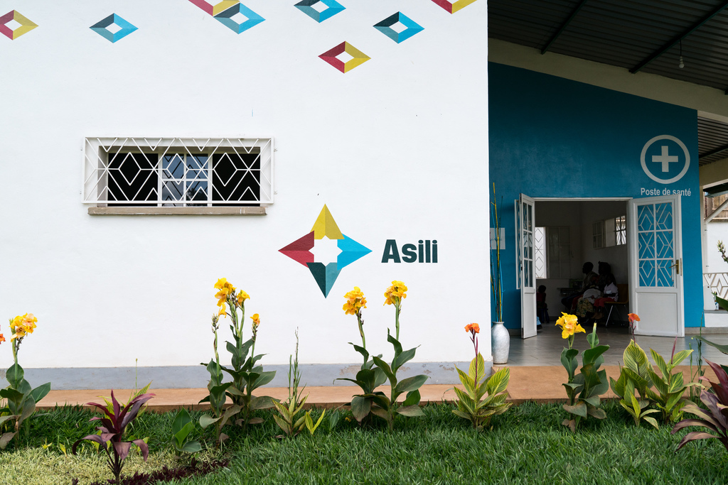 A white building, marked "Asili," with flowers growing in front, and a recessed porch to the right. 