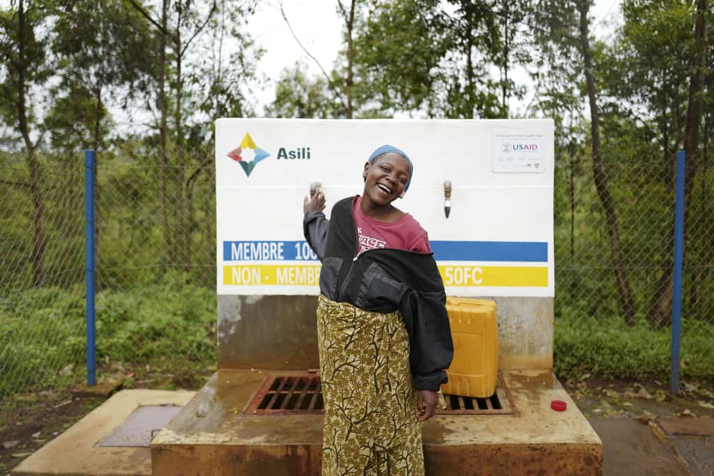 A woman laughs and points behind her to a cement water kiosk in a wooded area of Congo.